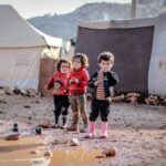 Children playing in a muddy refugee camp in Idlib, Syria, amidst tents and challenging conditions.