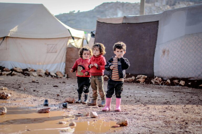 Children playing in a muddy refugee camp in Idlib, Syria, amidst tents and challenging conditions.