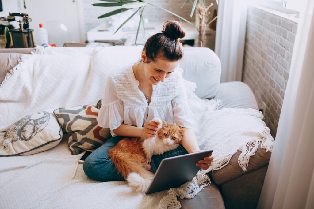 Young woman sitting on couch with her cat, browsing a tablet in a cozy home setting.