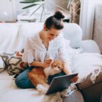 Young woman sitting on couch with her cat, browsing a tablet in a cozy home setting.